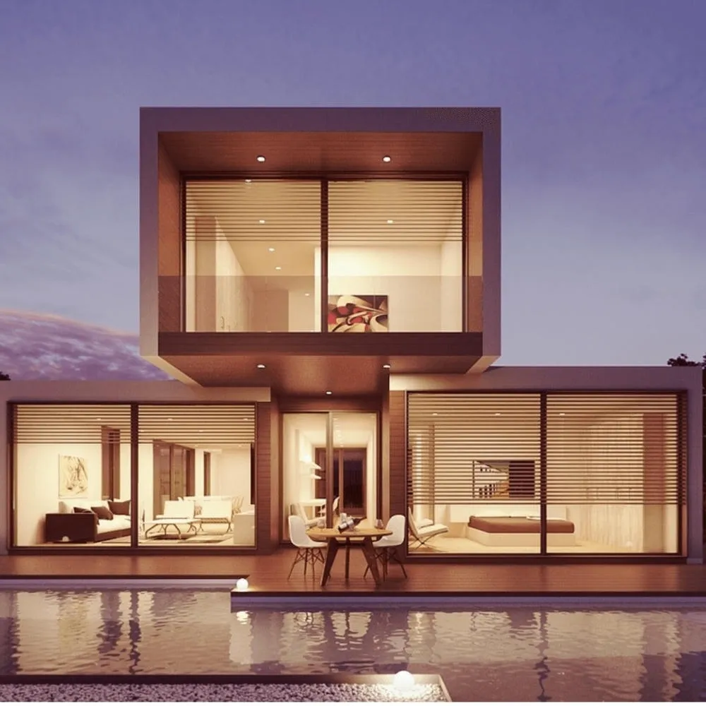 Modern modular house with cubic volumes, large windows, and a pool in the foreground, illuminated from within at dusk.