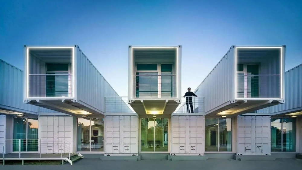 Multi-story buildings made of white prefabricated containers or modules, with illuminated balconies, under a blue sky at dusk.