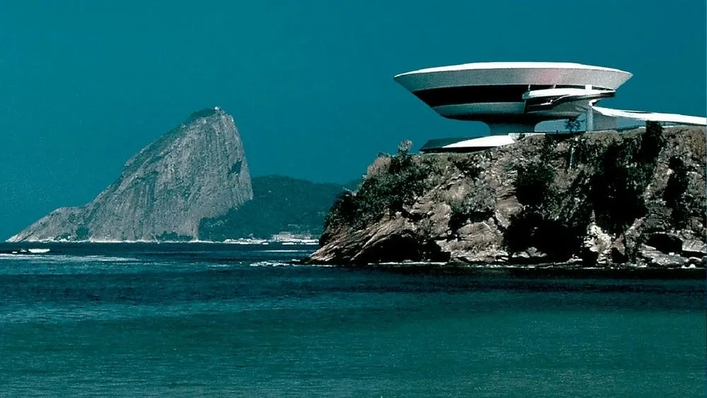 Vista do Museu de Arte Contemporânea de Niterói, com sua estrutura circular e futurista de concreto branco sobre uma falésia, em contraste com o mar e o Pão de Açúcar ao fundo, sob um céu azul-turquesa. O museu é um exemplo da arquitetura contemporânea no Brasil.