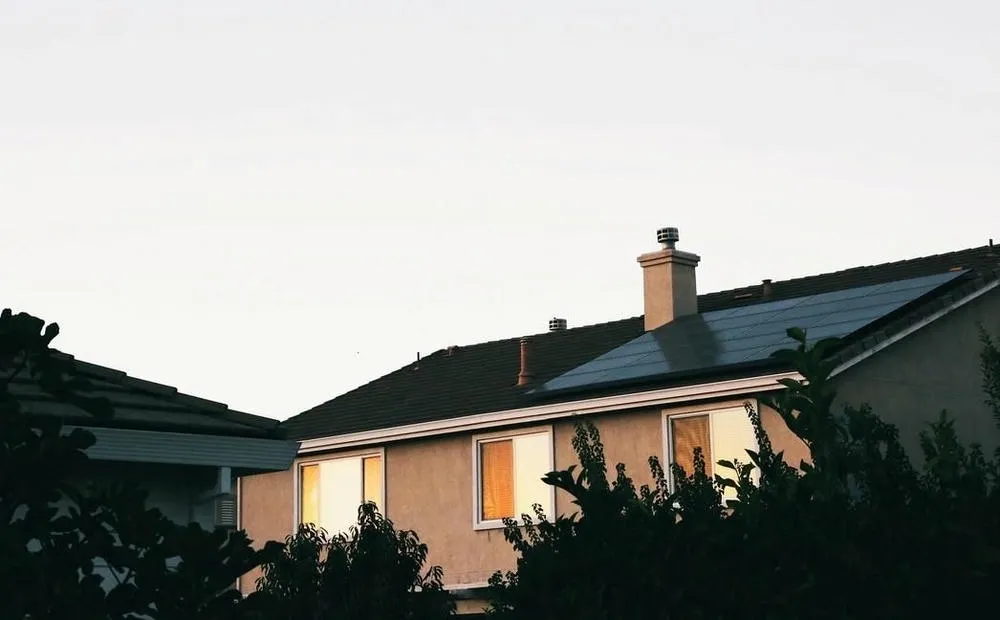 Uma casa com telhado escuro onde painéis solares estão instalados, visíveis acima de uma fileira de janelas que refletem a luz do pôr do sol, cercada por árvores escuras.