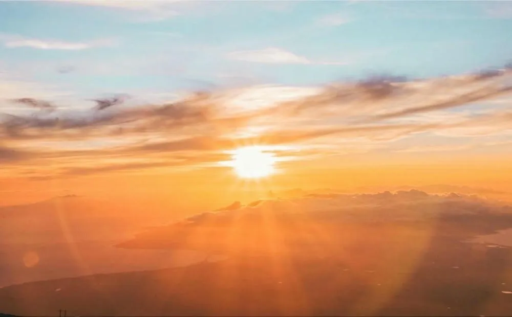 Um pôr do sol em tons de laranja e amarelo, com o sol brilhando intensamente sobre nuvens e uma paisagem costeira ao fundo. Raios de sol se espalham pelo céu.