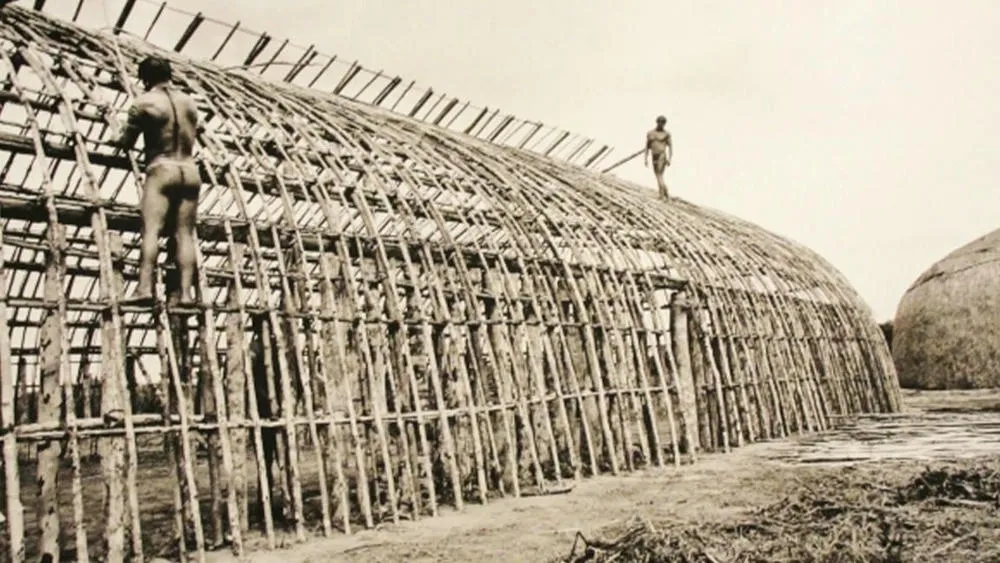 Representação da origem da arquitetura vernacular brasileira, com construções primitivas e materiais naturais.