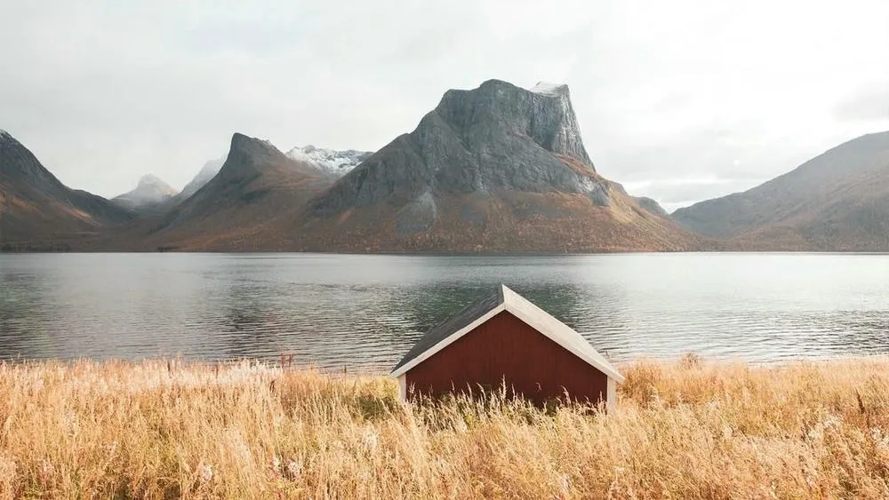 Uma casa pequena e tradicional com telhado vermelho em um campo de grama alta, com um lago e montanhas ao fundo, simbolizando a origem e a inspiração natural do estilo escandinavo.