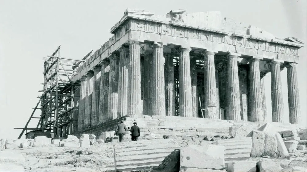 Vista em preto e branco do Partenon em Atenas, Grécia, com andaimes de madeira em uma das laterais, indicando um processo de restauração. A foto antiga realça a robustez das colunas dóricas e a grandiosidade do templo.