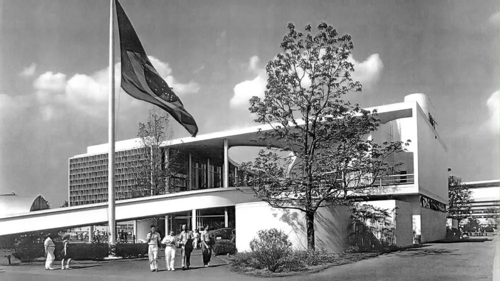 Antiga fotografia em preto e branco do Pavilhão Brasileiro na Feira Mundial de Nova York, projetado por Oscar Niemeyer e Lucio Costa, com a bandeira do Brasil hasteada, um exemplo da arquitetura futurista brasileira.