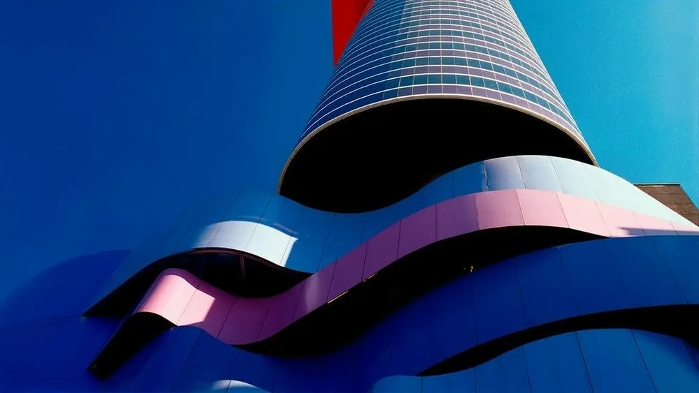 Vista inferior de um edifício de arquitetura contemporânea com formas curvas e abstratas em tons de azul e rosa, contrastando com um céu azul vibrante. Um pequeno elemento vermelho se projeta na parte superior do edifício.