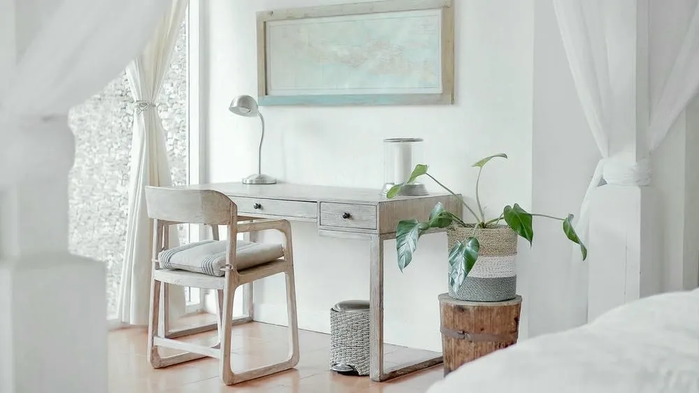 Um escritório minimalista em tons claros, com uma escrivaninha de madeira, cadeira, luminária de mesa, planta em vaso e um mapa emoldurado na parede, refletindo a simplicidade do design escandinavo.