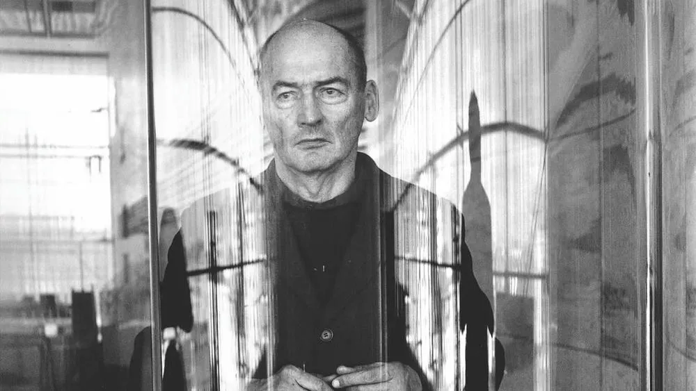 Retrato em preto e branco de Rem Koolhaas, arquiteto famoso, olhando para a frente através de uma superfície transparente.