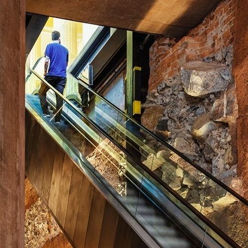 Vista de baixo para cima de uma escada rolante em um edifício com paredes de tijolos e pedras expostas e estrutura metálica, evidenciando o conceito de retrofit na arquitetura.