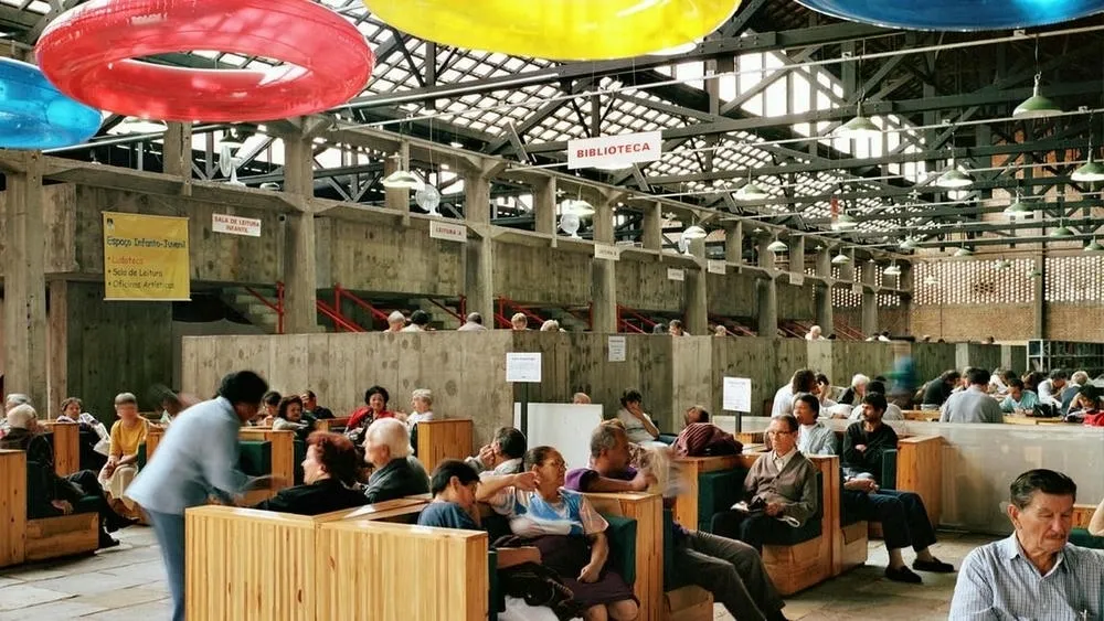 Interior of a large hall with concrete pillars, exposed ceiling, and people sitting on wooden benches, with large colorful rings hanging from the ceiling, representing the emergence of retrofit in architecture.