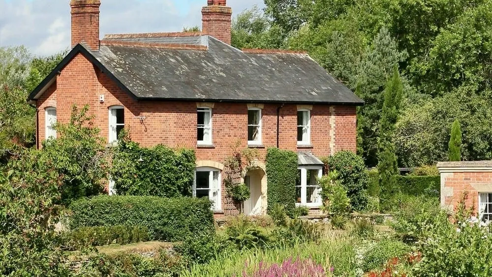 Rustic brick country house with a dark roof, surrounded by trees and a flower garden, conveying tranquility.