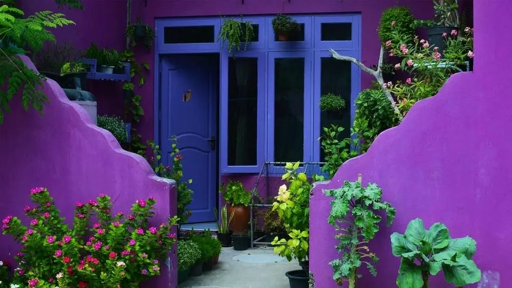 Uma sala de estar com pé-direito duplo, grandes janelas e um sofá cinza com almofadas azuis. O ambiente é amplo e iluminado, com tons frios predominantes nas paredes e mobiliário, que ampliam a percepção do espaço.