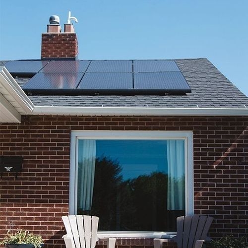 Facade of a brick house with a dark roof, where solar panels are installed. A large window reflects the blue sky, and white chairs are on the front patio.