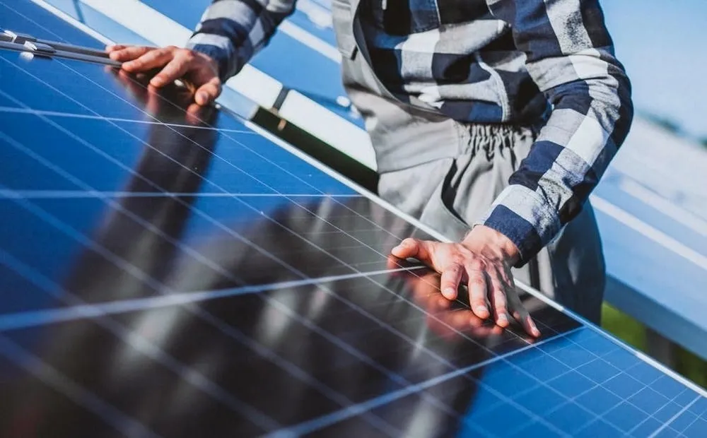 Person installing a solar panel on a roof, with hands visible on the dark surface of the panel, reflecting the blue sky. The person is wearing a plaid shirt and overalls.