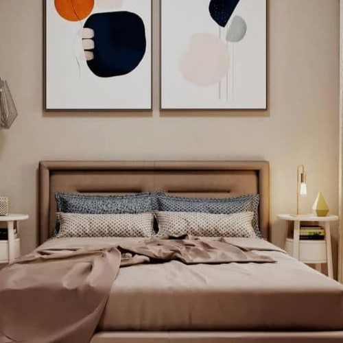 Detalhe de uma cama de casal com cabeceira estofada, lençóis neutros e travesseiros com estampas geométricas e azuis. Acima da cama, dois quadros com arte abstrata em tons de laranja, azul marinho, rosa e cinza, emoldurados em branco. Uma luminária de mesa dourada e um abajur geométrico descansam sobre uma mesa de cabeceira branca.
