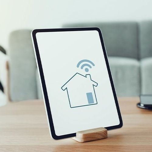 Tablet on a wooden stand displaying a house icon with a Wi-Fi signal, representing home automation. In the background, a sofa and a plant, in a well-lit environment.