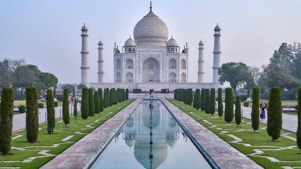 Vista do Taj Mahal, na Índia, com sua estrutura branca refletida em um longo espelho d'água, realçando a simetria.