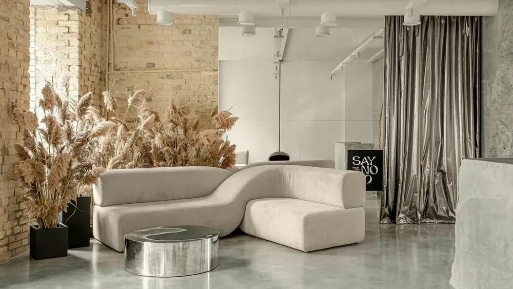 Sala de estar com sofá curvo em tom neutro, parede de tijolos expostos e folhagens secas, destacando o uso de cores neutras e texturas naturais na decoração.