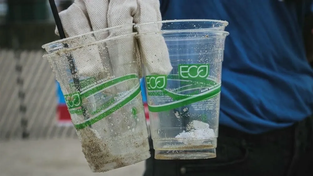 Persona sosteniendo dos vasos de plástico desechables con etiquetas 'eco', simbolizando el greenwashing.