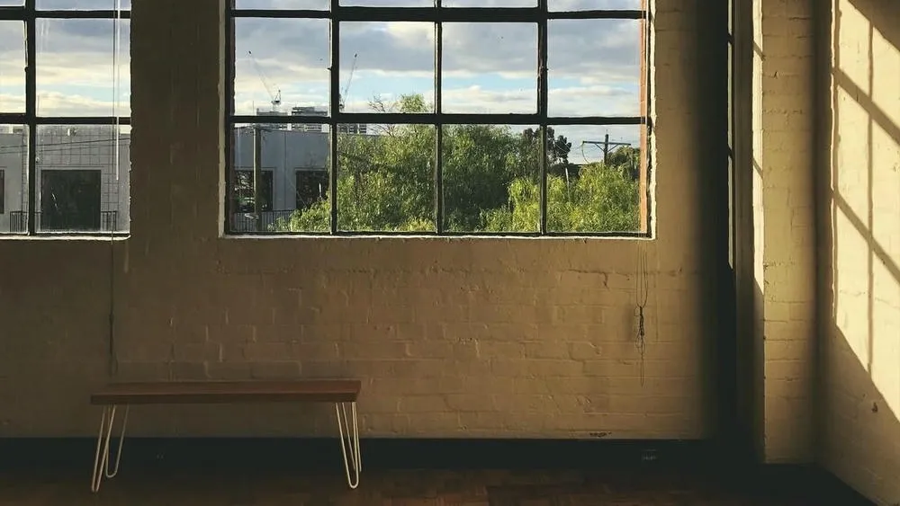Pared con dos ventanas amplias que permiten la entrada de luz solar, creando un fuerte contraste de sombra. Un banco de madera minimalista está posicionado debajo de la ventana, y la pared exhibe una textura rústica.