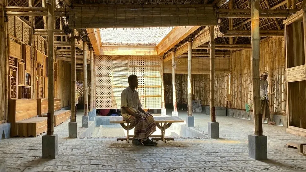 Man sitting on a wooden bench inside a bamboo and wood structure, with natural light entering through the roof.