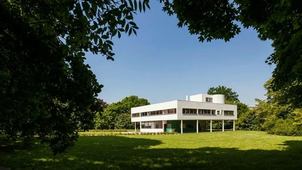 View of Villa Savoye on a sunny day, with its white facade, horizontal windows, and pilotis, surrounded by green grass and trees.