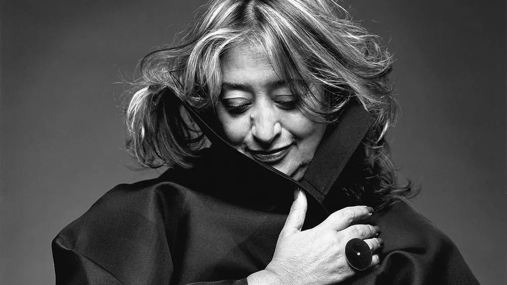 Black and white portrait of Zaha Hadid, with gray hair and holding her coat collar.